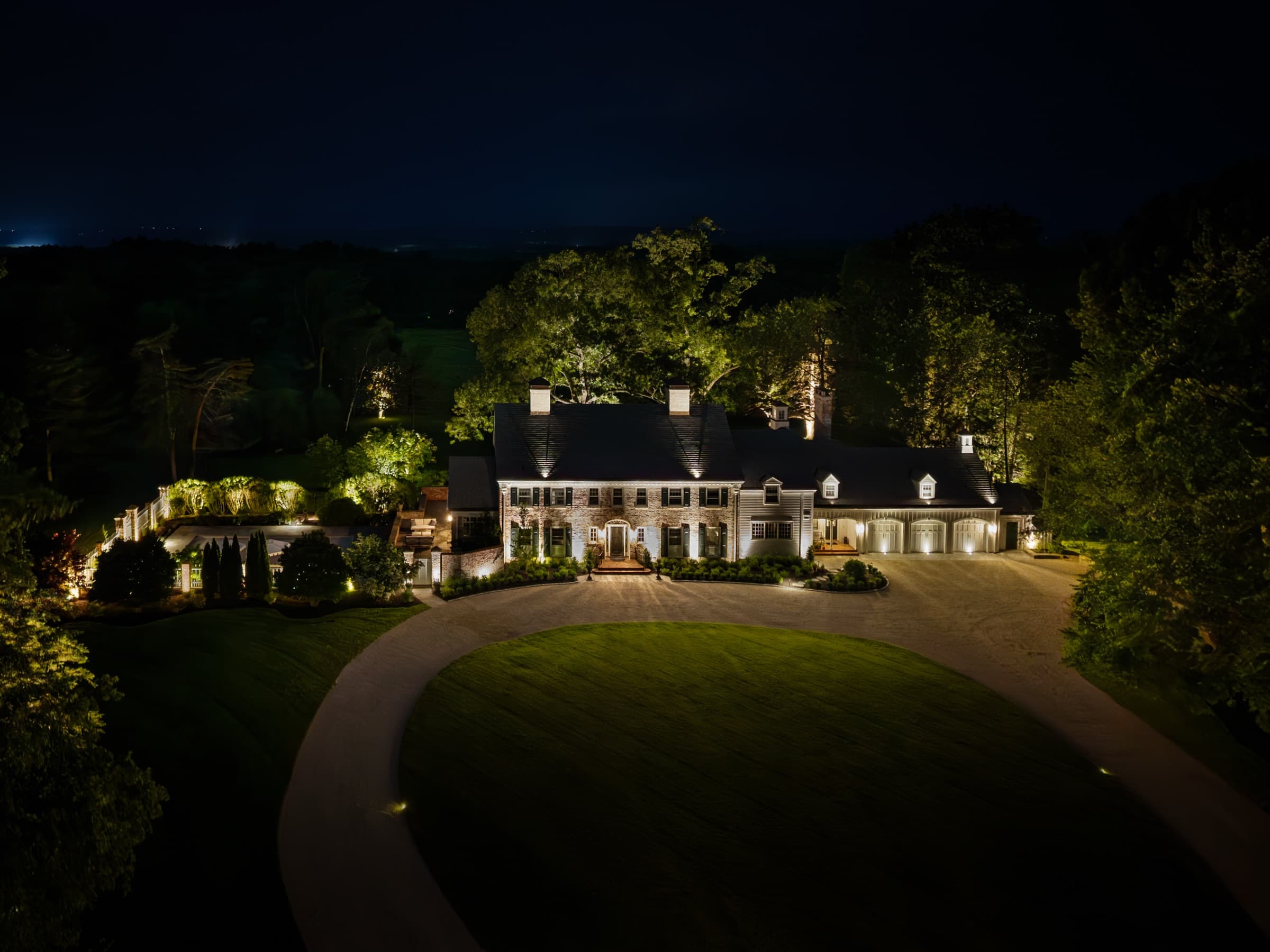 Aerial view at blue hour: lit façade, circular lawn and terraced garden beyond
