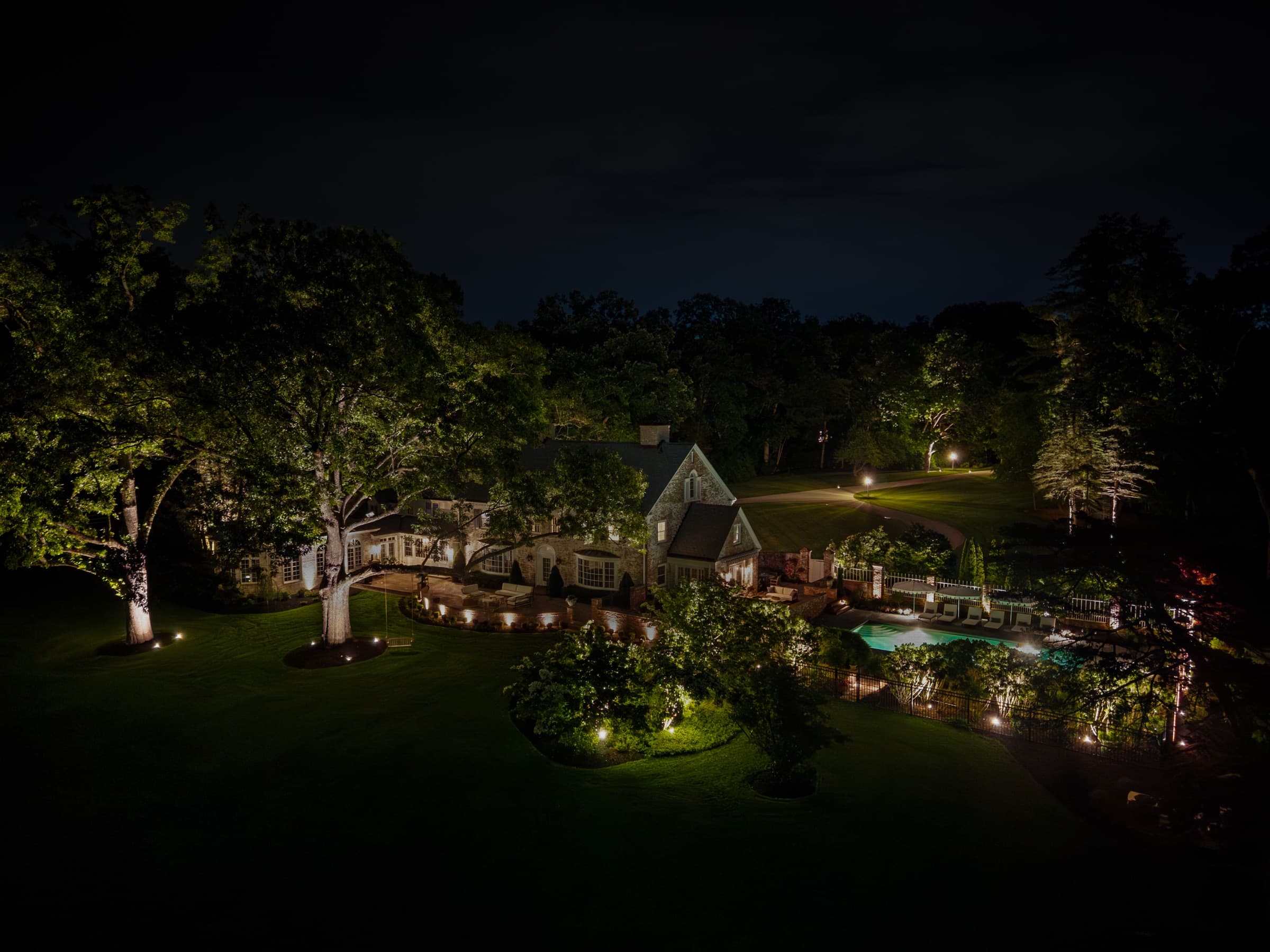The lit house and grounds seen from the air, drive curving in from the left