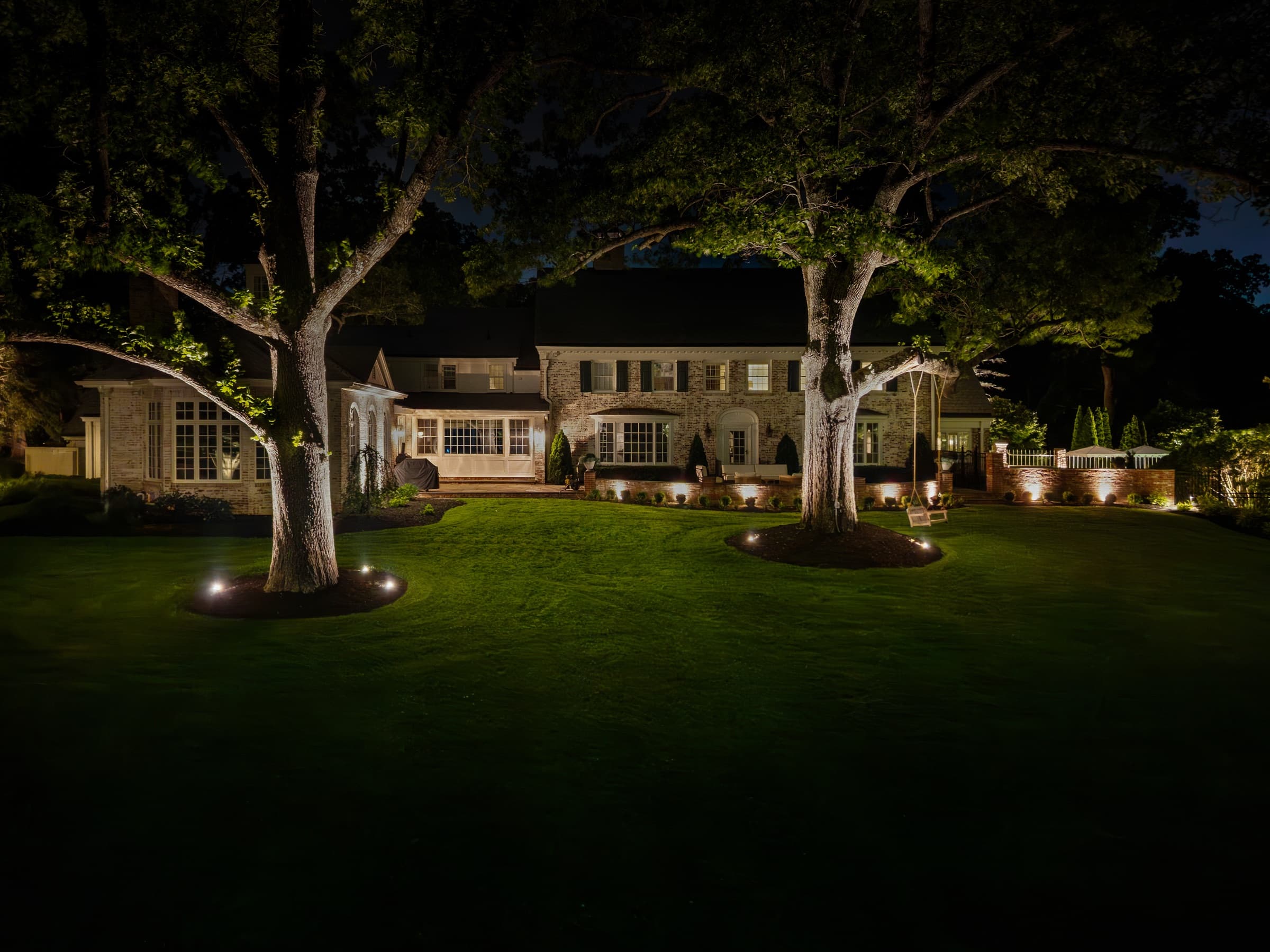 Uplit specimen trees over a lawn at night, with the house beyond