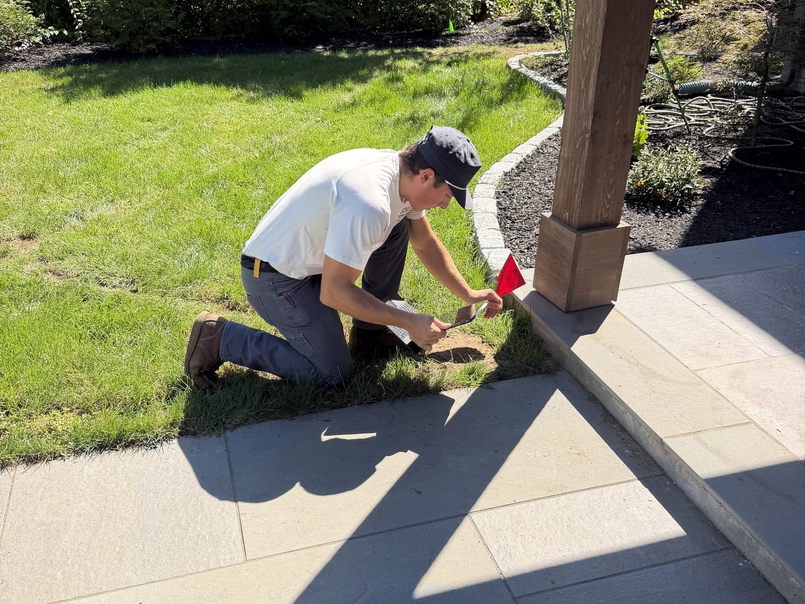 Kneeling on the lawn to place a red marker flag beside a porch post